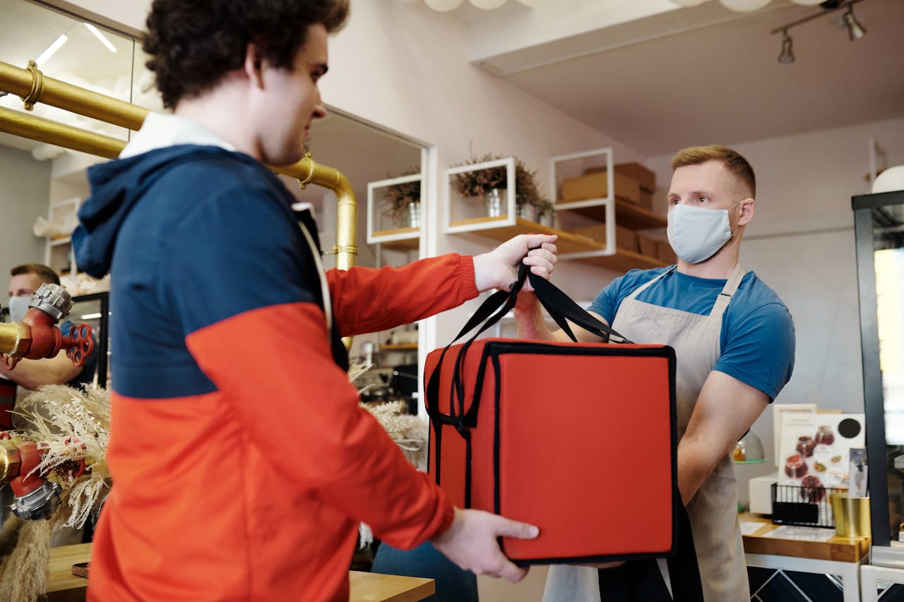 Two men exchanging a food delivery bag indoors, both wearing face masks.